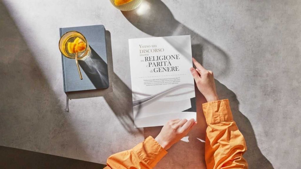 Mani femminili tengono una pubblicazione Bah&aacute;&rsquo;&iacute; intitolata &ldquo;Verso un discorso nuovo su religione e parit&agrave; di genere&rdquo; accanto a un taccuino e un bicchiere con fette d&rsquo;arancia su un tavolo illuminato dalla luce naturale